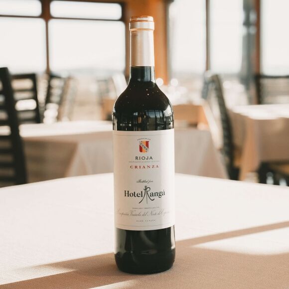 A bottle of CVNE Rioja Crianza red wine, custom bottled for Hotel Rangá, standing on a white tablecloth in a brightly lit dining room with blurred seating in the background.