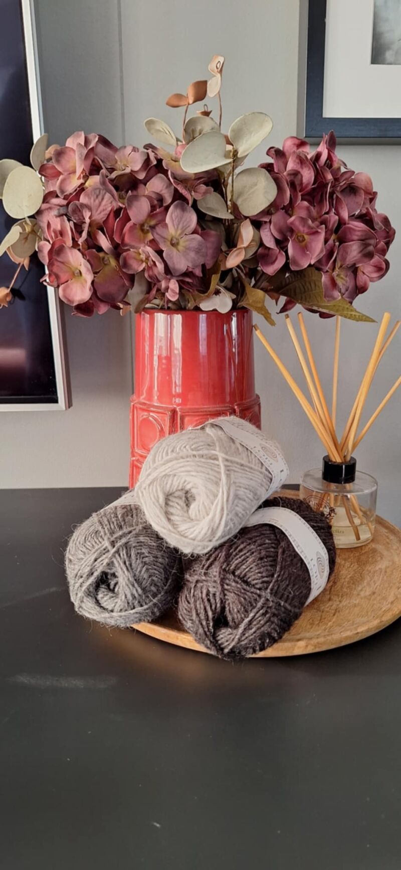 A photo of three skeins of grey yarn in varying shades beside a vase filled with purple flowers.