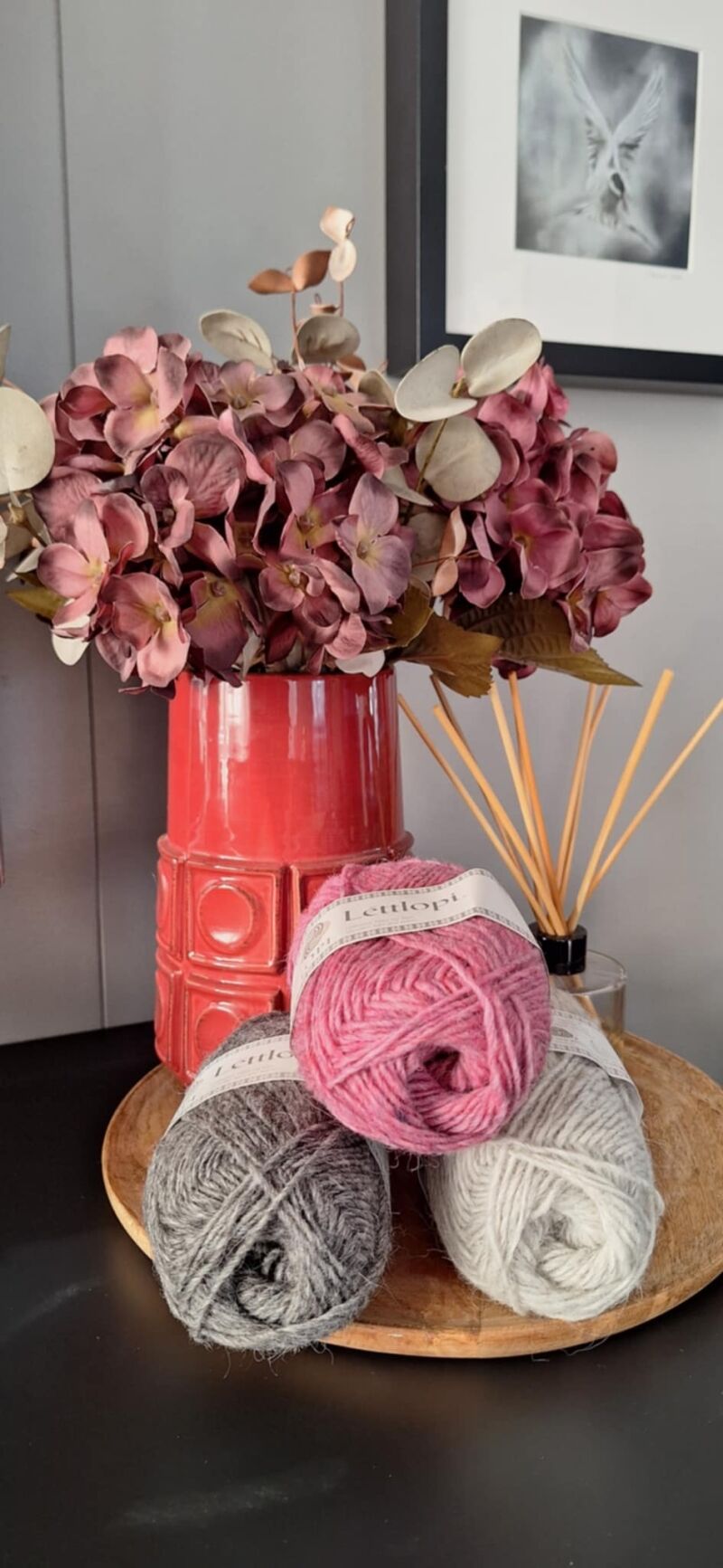 A photo of three skeins of yarn in varying shades beside a vase filled with purple flowers.