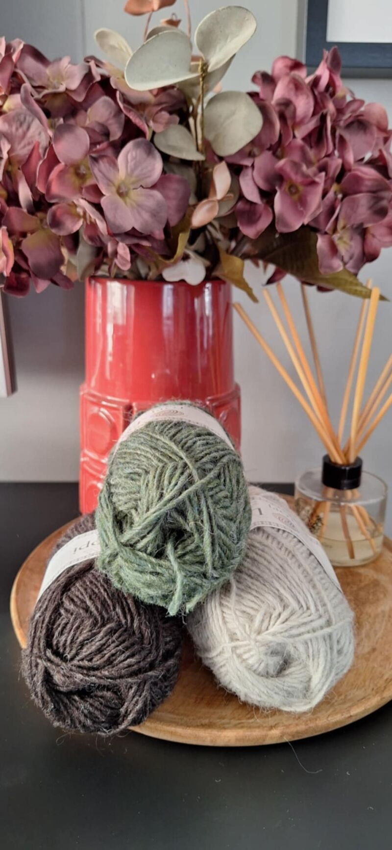 A photo of three skeins of yarn in varying shades beside a vase filled with purple flowers.
