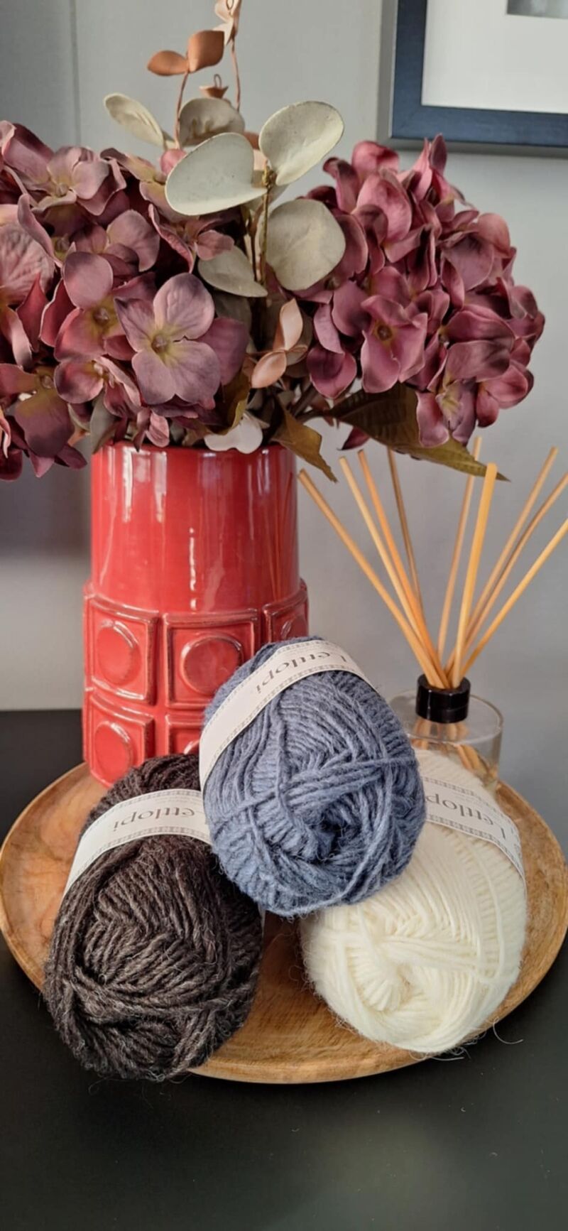 A photo of three skeins of yarn in varying shades beside a vase filled with purple flowers.