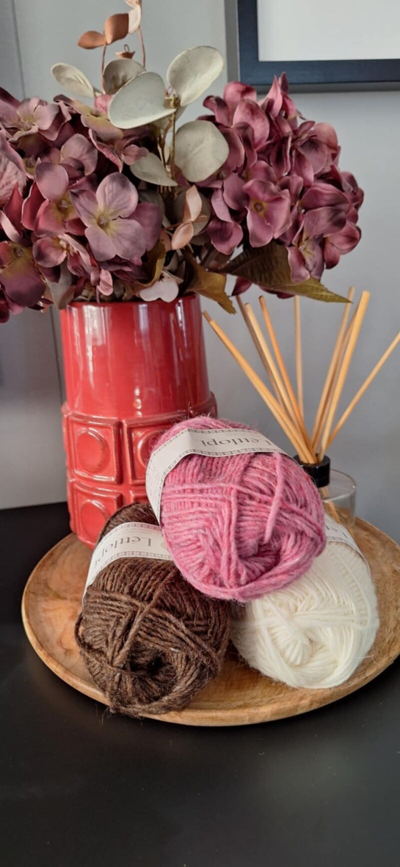 A photo of three skeins of yarn in varying shades beside a vase filled with purple flowers.