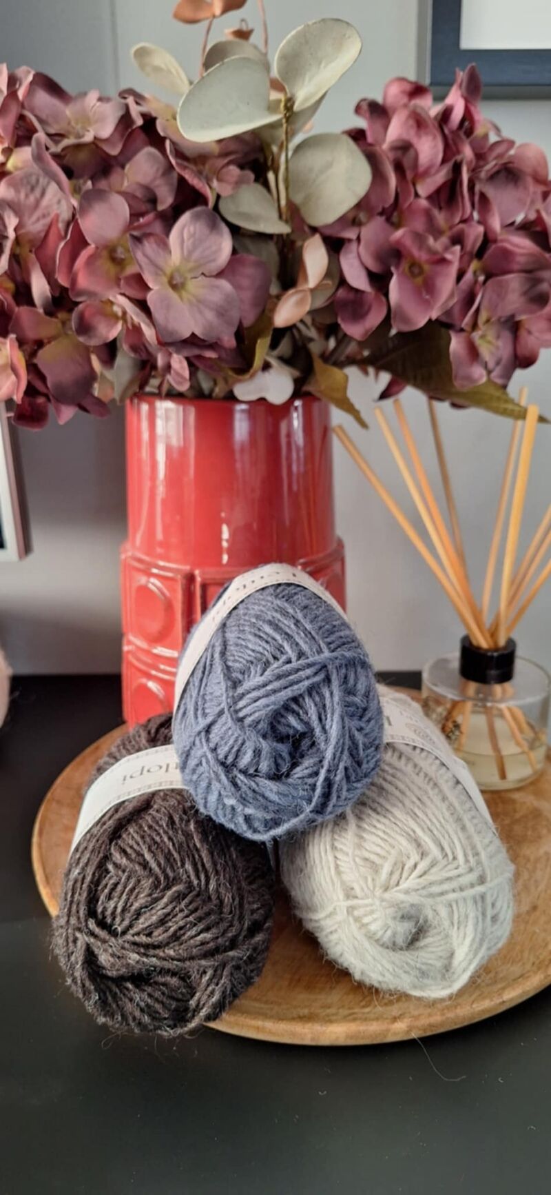 A photo of three skeins of yarn in varying shades beside a vase filled with purple flowers.