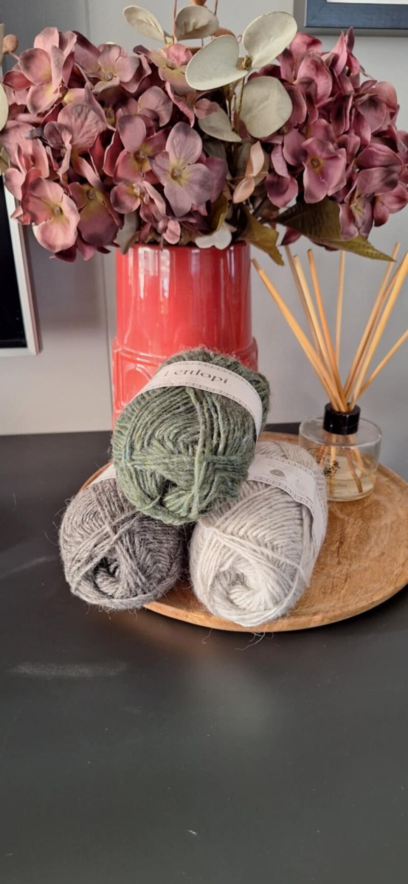 A photo of three skeins of yarn in varying shades beside a vase filled with purple flowers.
