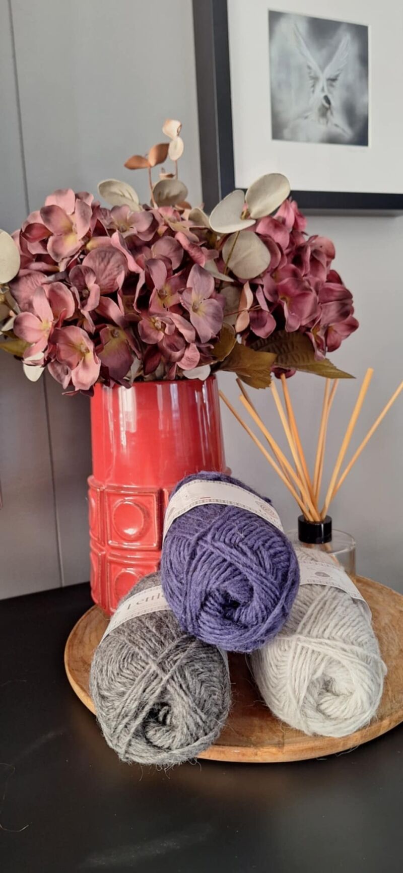 A photo of three skeins of yarn in varying shades beside a vase filled with purple flowers.