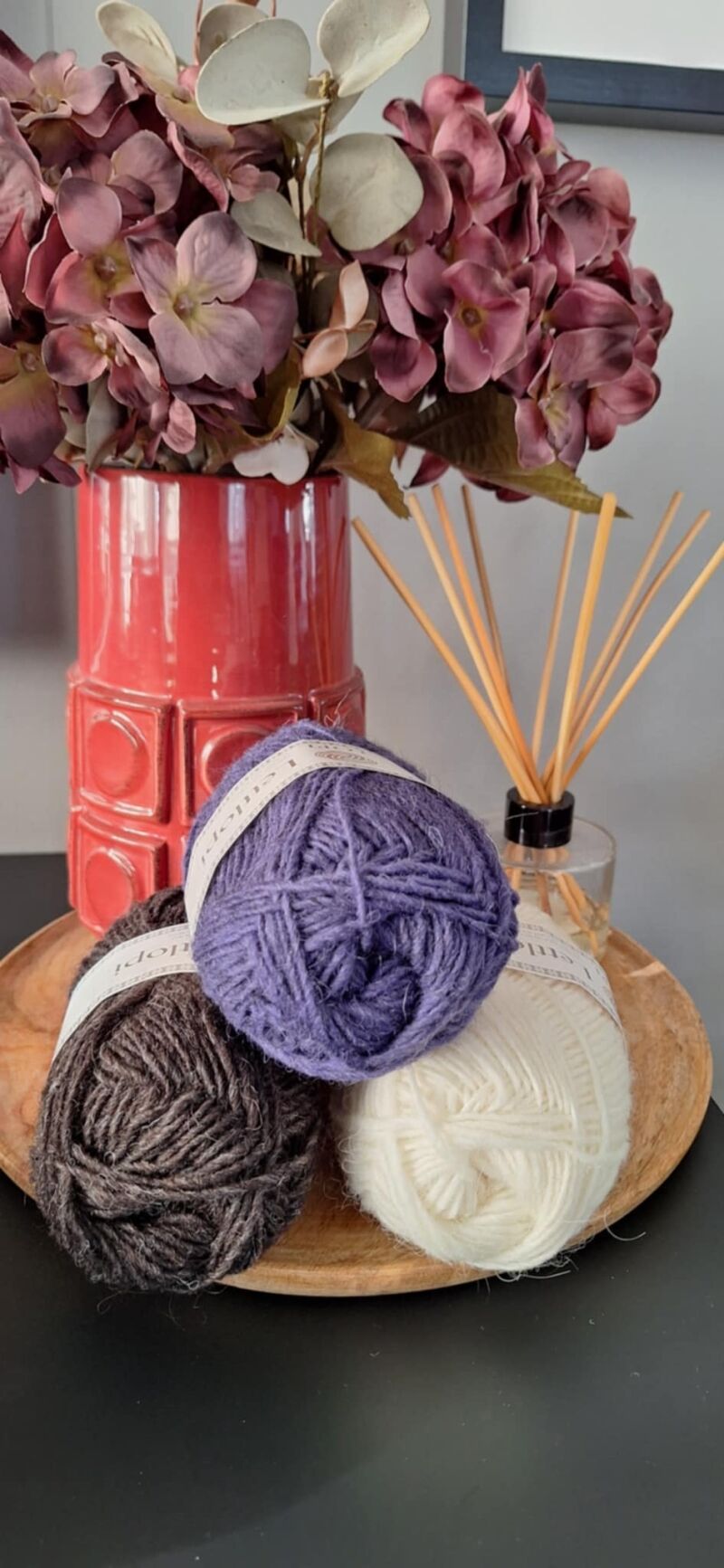 A photo of three skeins of yarn in varying shades beside a vase filled with purple flowers.