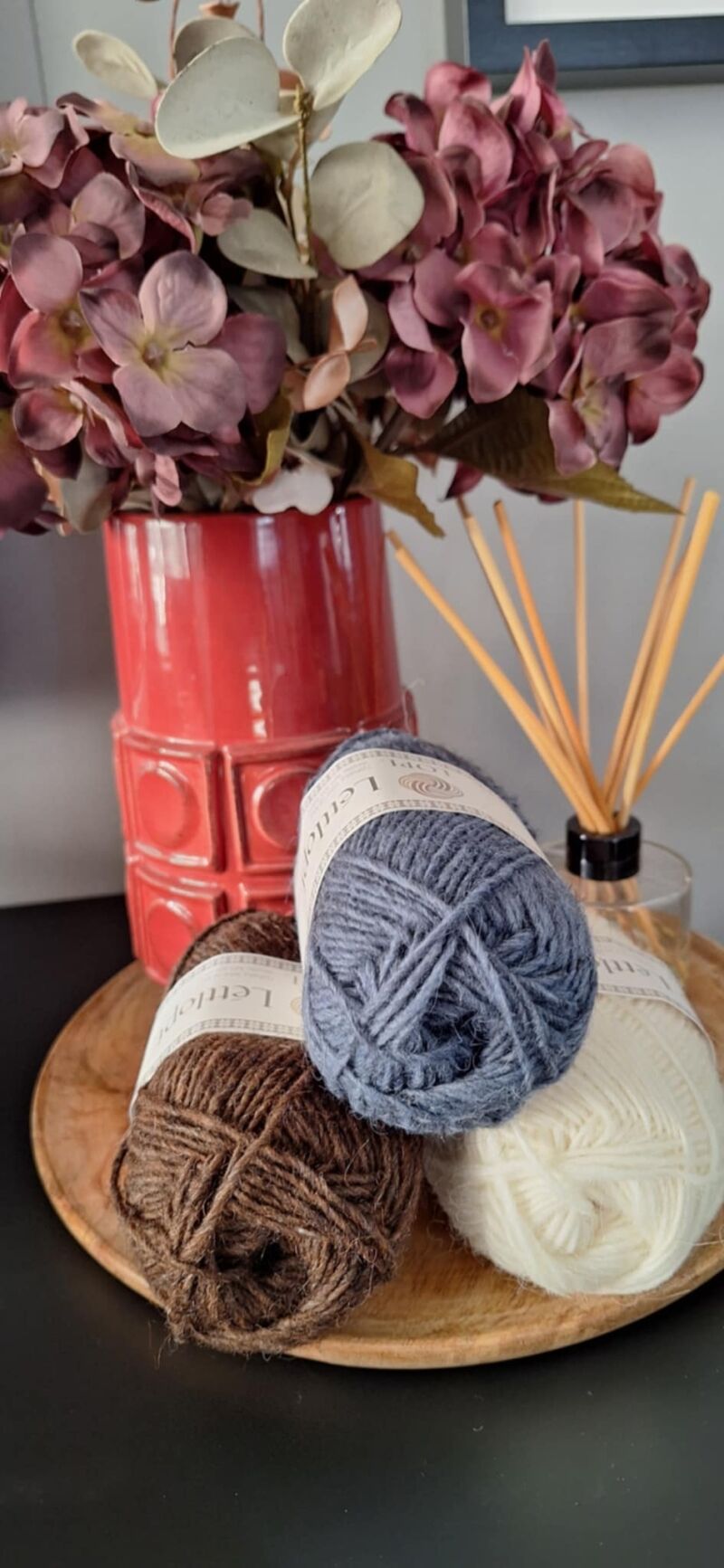 A photo of three skeins of yarn in varying shades beside a vase filled with purple flowers.
