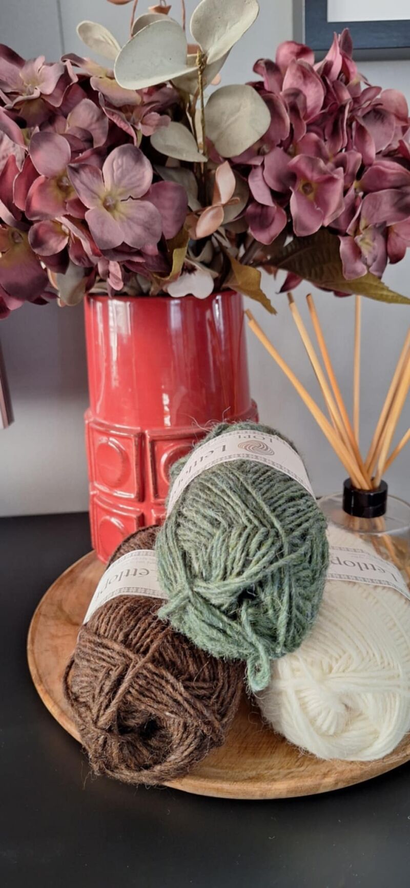 A photo of three skeins of yarn in varying shades beside a vase filled with purple flowers.