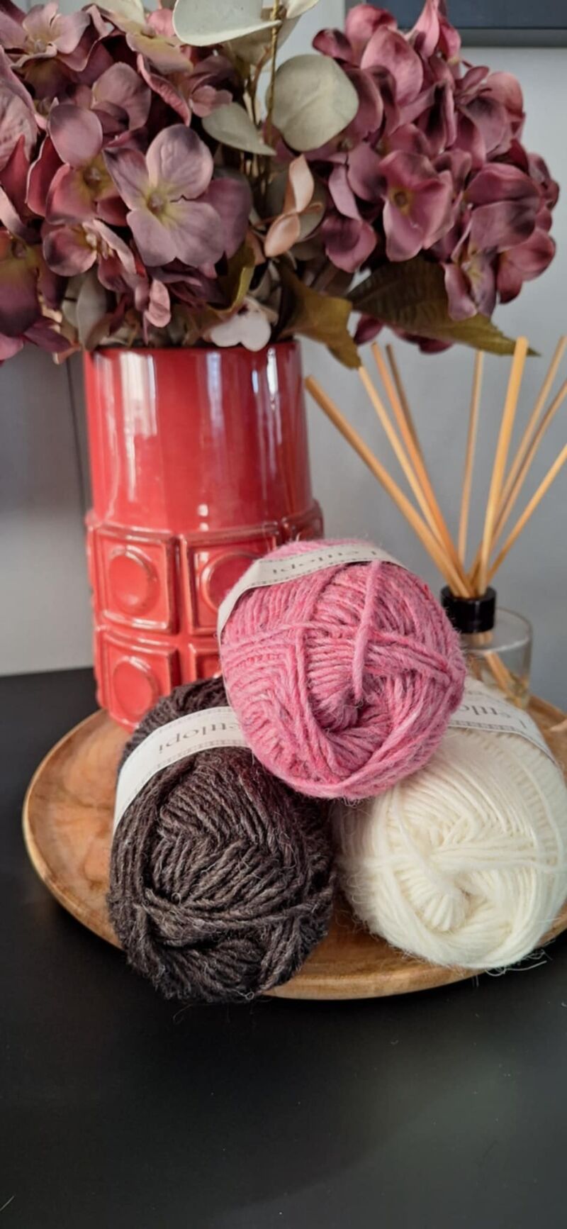 A photo of three skeins of yarn in varying shades beside a vase filled with purple flowers.